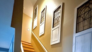 Una escalera de madera con barandas blancas, flanqueada por paredes con marcos de ventana decorativos y una planta en maceta.