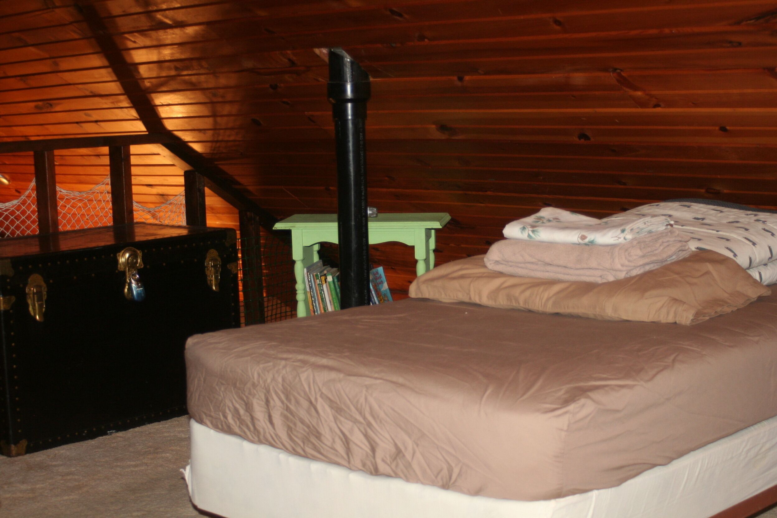 Une chambre avec un plafond en bois, un lit recouvert d’une couverture beige, une table de chevet verte avec des livres et une valise noire.