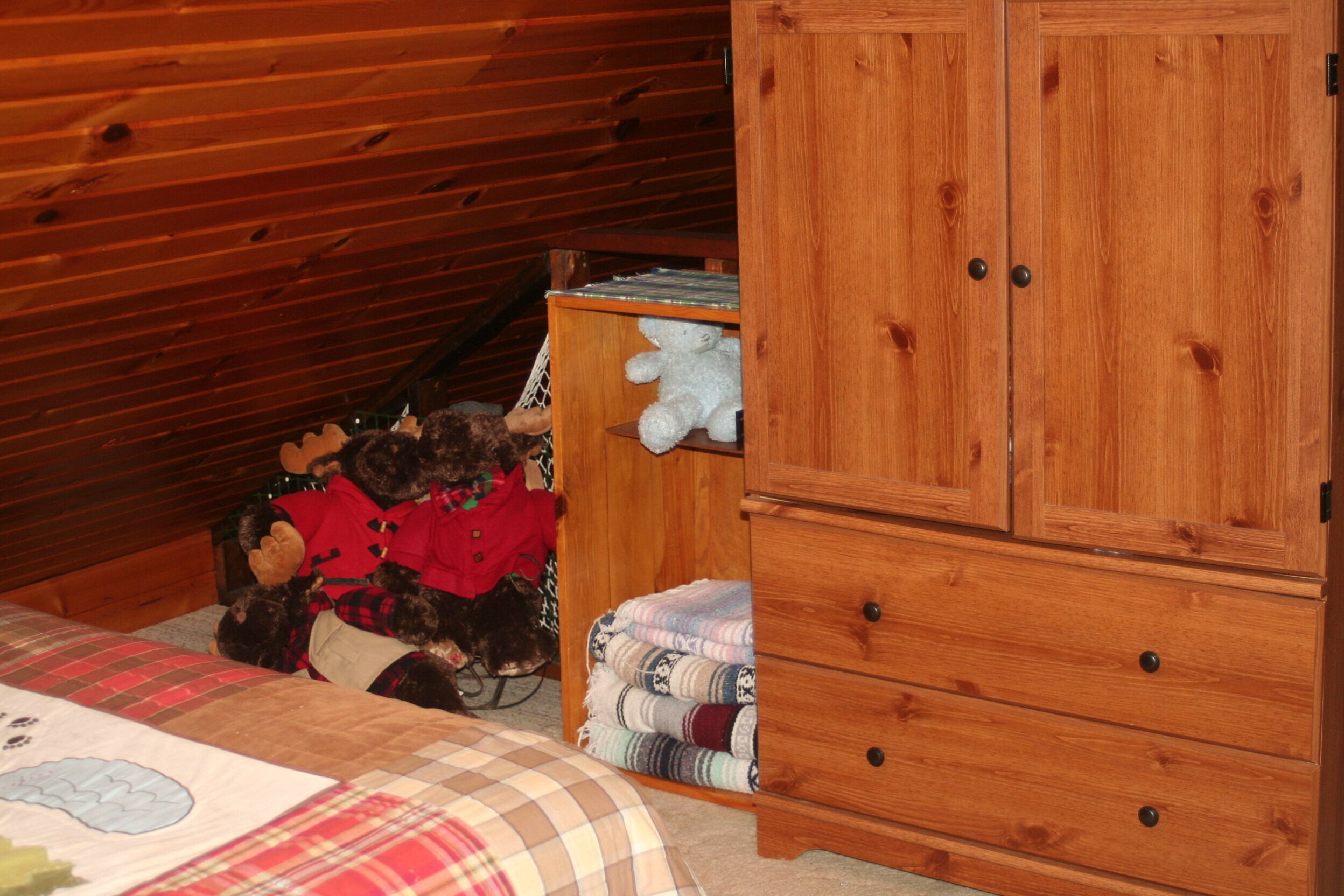 Ein Schlafzimmer mit Holzboden, einem Bett mit kariertem Überwurf, einem Holzschrank und einem Regal mit Stofftieren und gefalteten Decken.