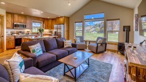 A spacious living area with a dark brown sofa, a coffee table, and a fireplace. There is a kitchen with wooden cabinets and a microwave. Large windows provide a view of the outdoors.