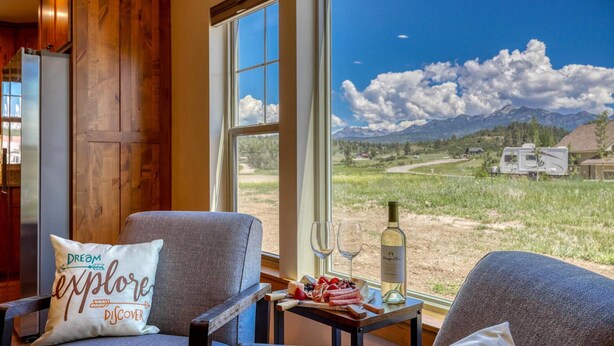 A room with a large window offering a view of a mountainous landscape, a dining table with a bottle of wine, and two chairs with a cushion that reads "Dream Explore Discover".