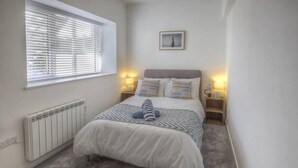A bedroom with a bed, a radiator, a window with blinds, a bedside table with a lamp, and a picture on the wall. - The Swell by Guestz (Rhosneigr)