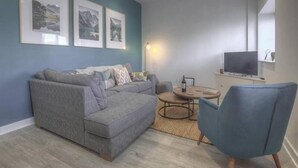 A living room with a grey sectional sofa, a round coffee table, and a blue armchair. There are framed pictures on the wall and a flat-screen TV mounted above a white cabinet. - The Swell by Guestz (Rhosneigr)