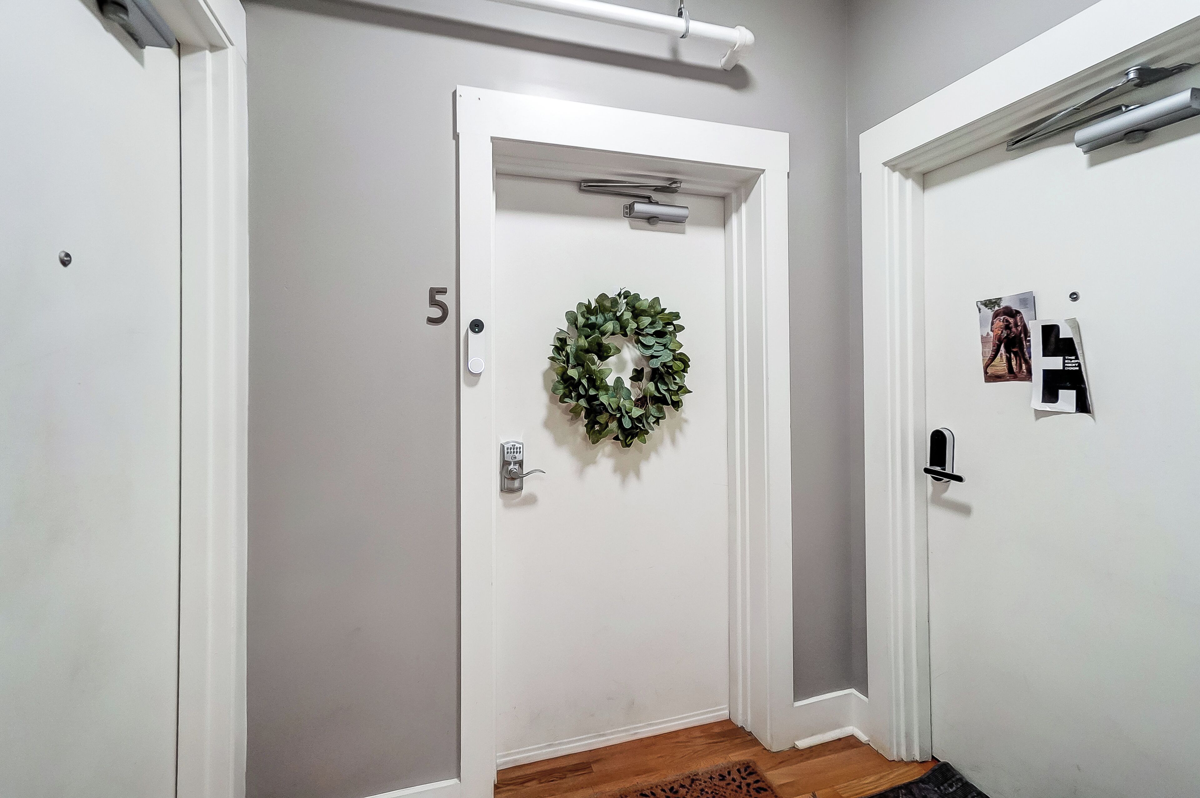 A hallway with two white doors, one with a wreath and the other with a photo and a black object.