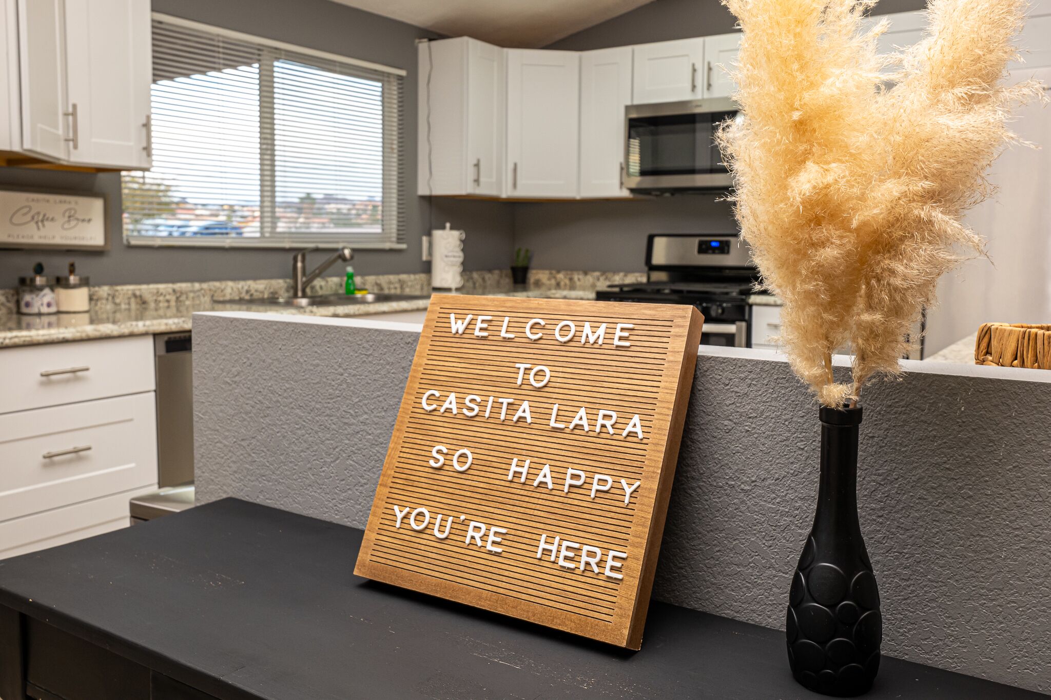 A kitchen with a welcome sign, a black vase with pampas grass, and a view of the city through the window.