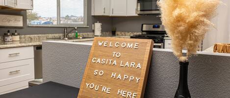 A kitchen with a welcome sign, a black vase with pampas grass, and a view of the city through the window.