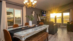Een eetgedeelte met een glazen tafel, stoelen, een bank, een kroonluchter, een potplant en een televisie aan de muur.