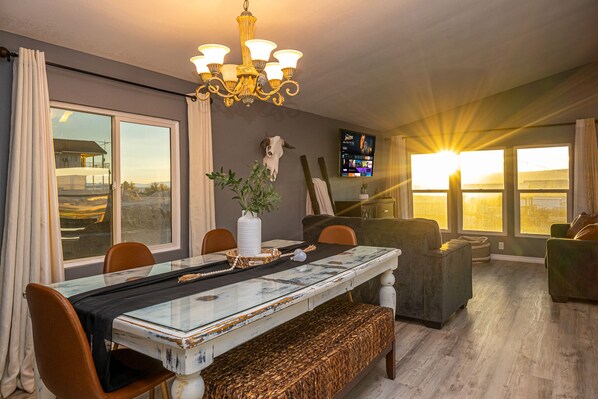A dining area with a glass table, chairs, a bench, a chandelier, a potted plant, and a TV on the wall.