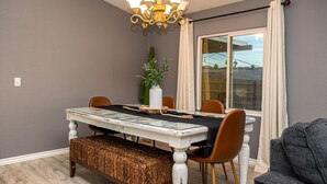 Een eetgedeelte met een tafel met glazen blad, houten stoelen, een bank, een kroonluchter en een groot raam met gordijnen.