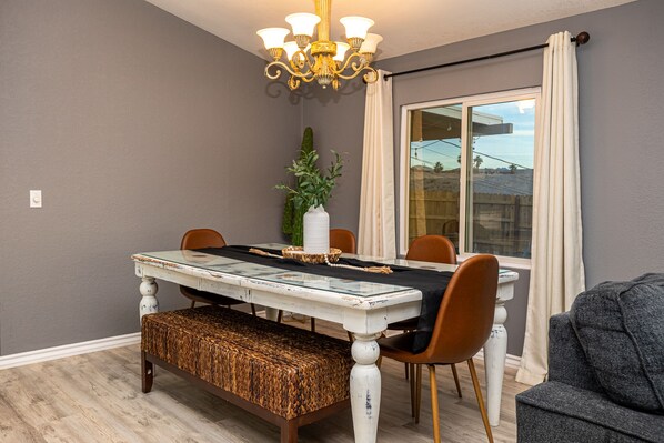 A dining area with a glass-top table, wooden chairs, a bench, a chandelier, and a large window with curtains.