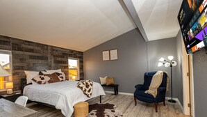Ein modernes Schlafzimmer mit Bett, Nachttisch, Lampe, Stuhl und einem großen Fernseher.
