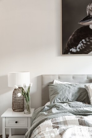 Een net opgemaakt bed met een grijs-witte gewatteerde dekbedovertrek, een wit nachtkastje met een lamp en een vaas tulpen, en een ingelijst beeld op de muur.