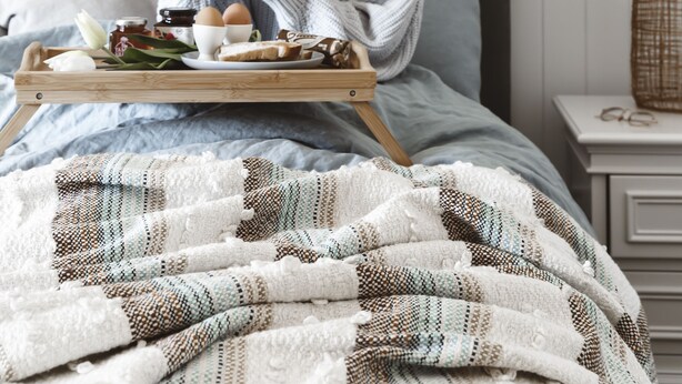 Ein ordentlich bezogenes Bett mit grauem Kopfteil, ein Holztablett mit Frühstücksgegenständen und ein Holzschläger, der an der Wand lehnt.