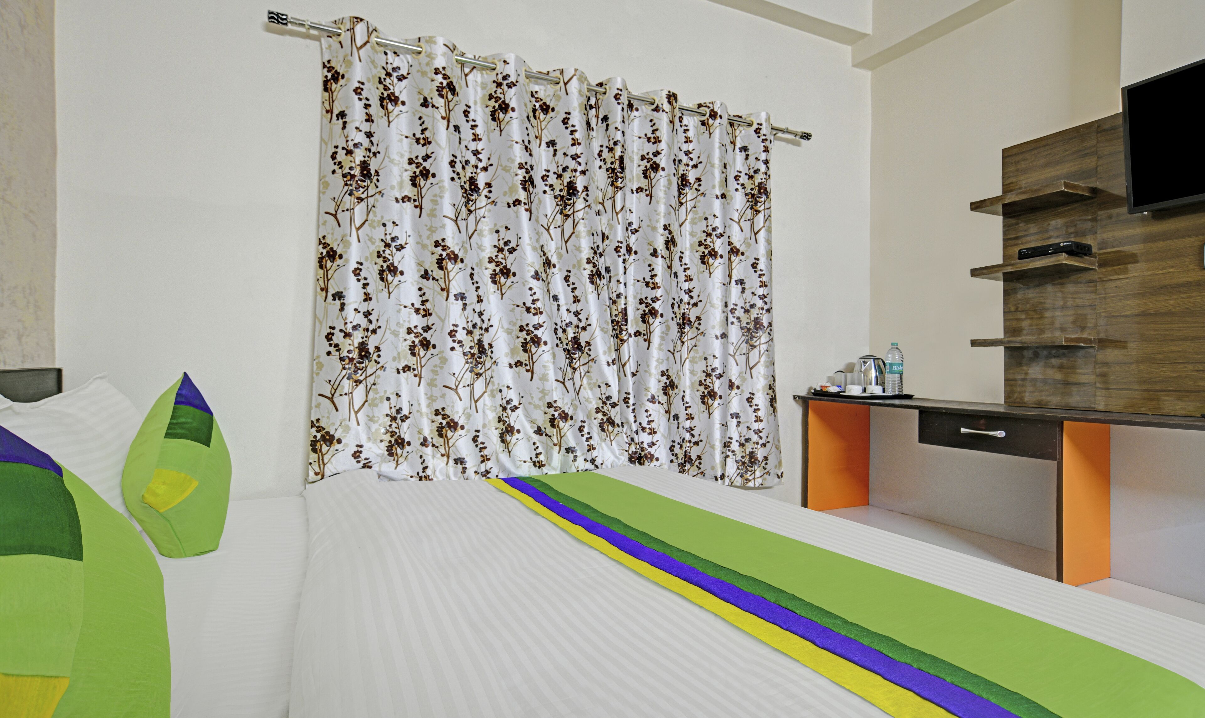 Moderni hotellihuone, jossa on sänky, kukkapainokuviollinen suihkuverho, puinen meikkipöytä peilillä sekä seinälle asennettu litteänäyttöinen televisio.