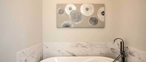 A bathroom with a freestanding bathtub, marble tiles, and a wall-mounted abstract art piece.