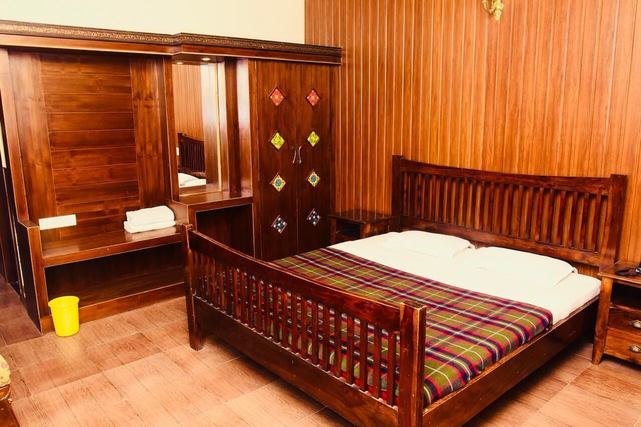Een houten hemelbed met een kleurrijke geruite plaid, een houten garderobe met decoratieve diamantmotieven, en een houten nachtkastje met een lamp.