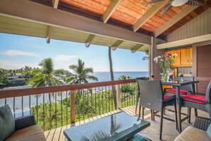 A balcony with wicker furniture, a glass table, and a view of the ocean and palm trees.