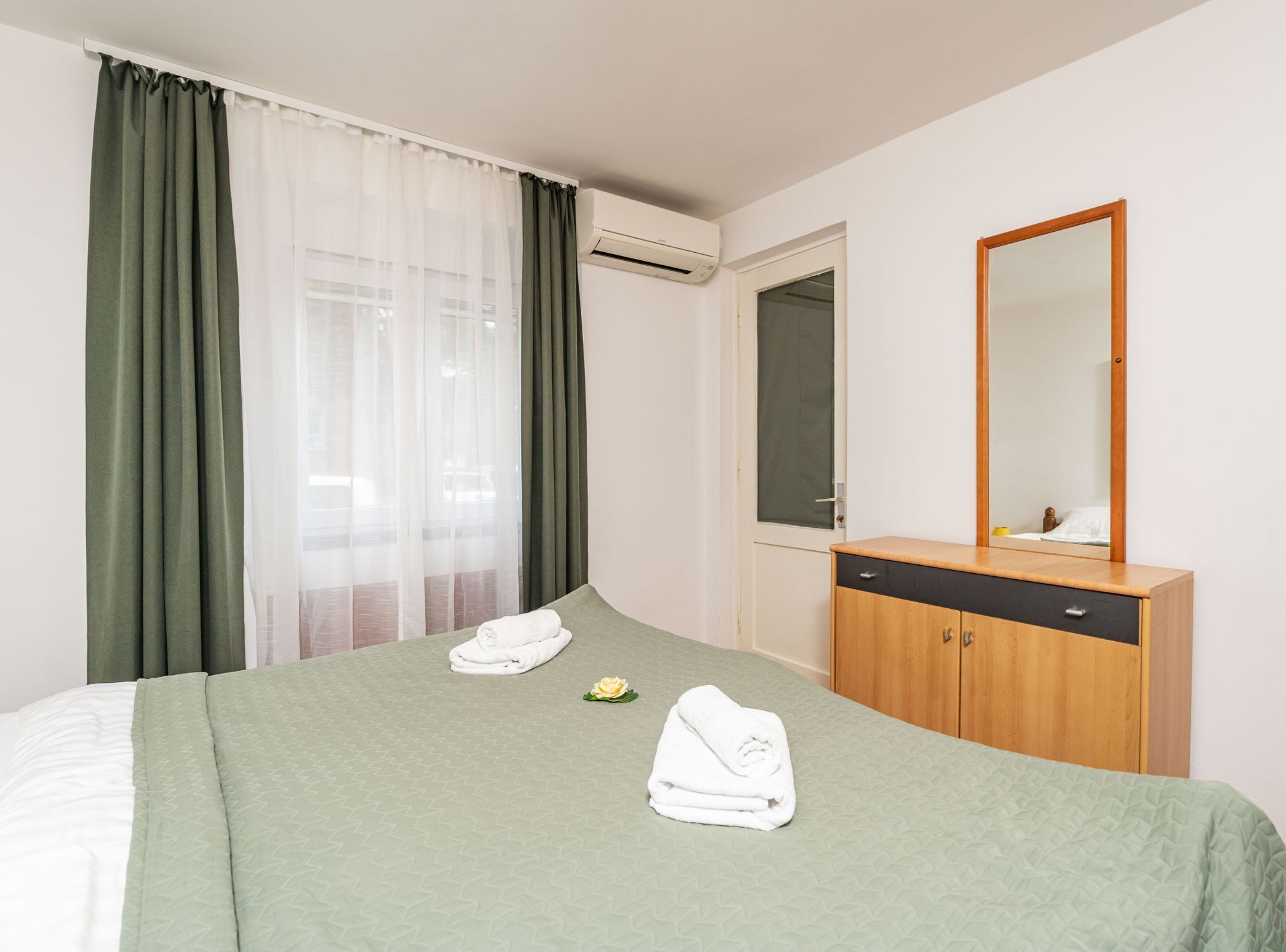 Une chambre d’hôtel avec un lit, une commode en bois, un miroir et une fenêtre avec des rideaux.