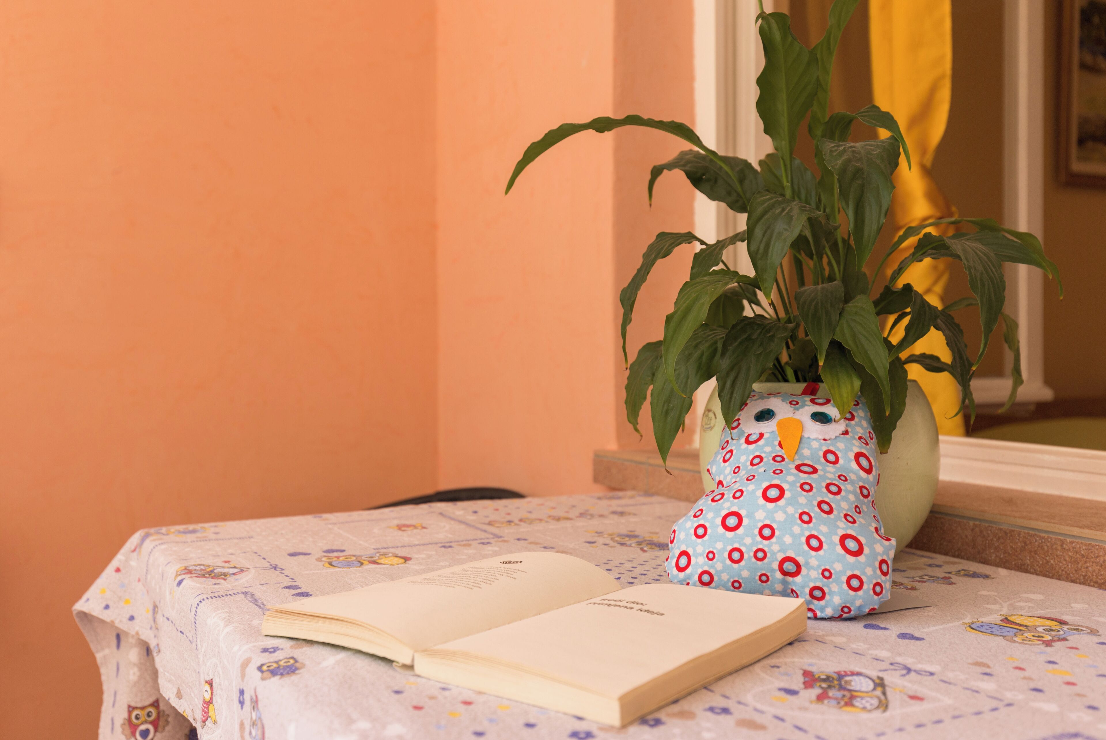 Un libro abierto sobre una mesa con mantel estampado, una planta en maceta en un jarrón decorativo y una cortina amarilla al fondo.
