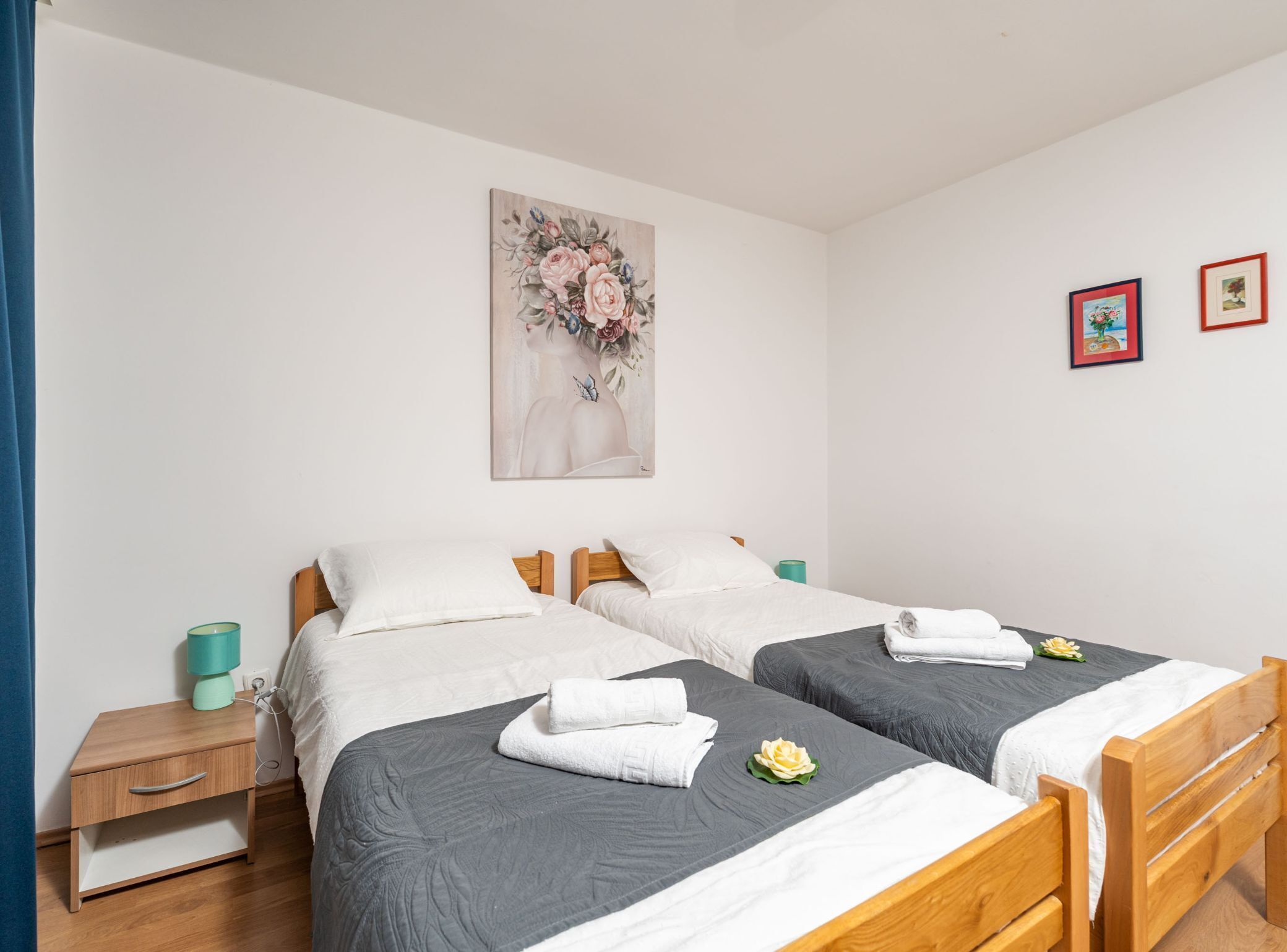 Ein Zimmer mit zwei Betten, jedes mit einer grauen Bettdecke und weißen Kissen. Ein türkisblaues Nachttischchen mit einer Lampe und ein Blumenbild an der Wand sind ebenfalls vorhanden.