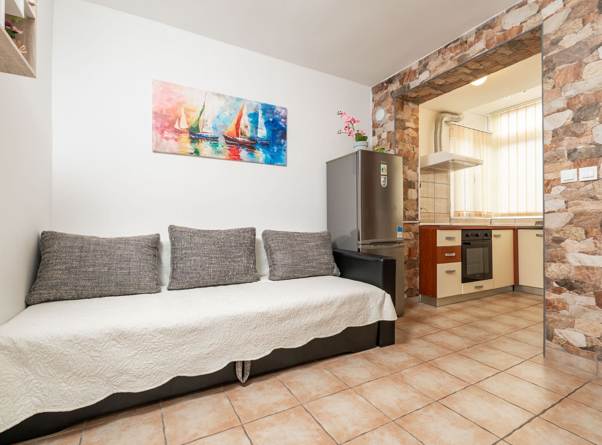 A cozy living area with a sofa, a painting, a refrigerator, and a kitchen with stone walls.