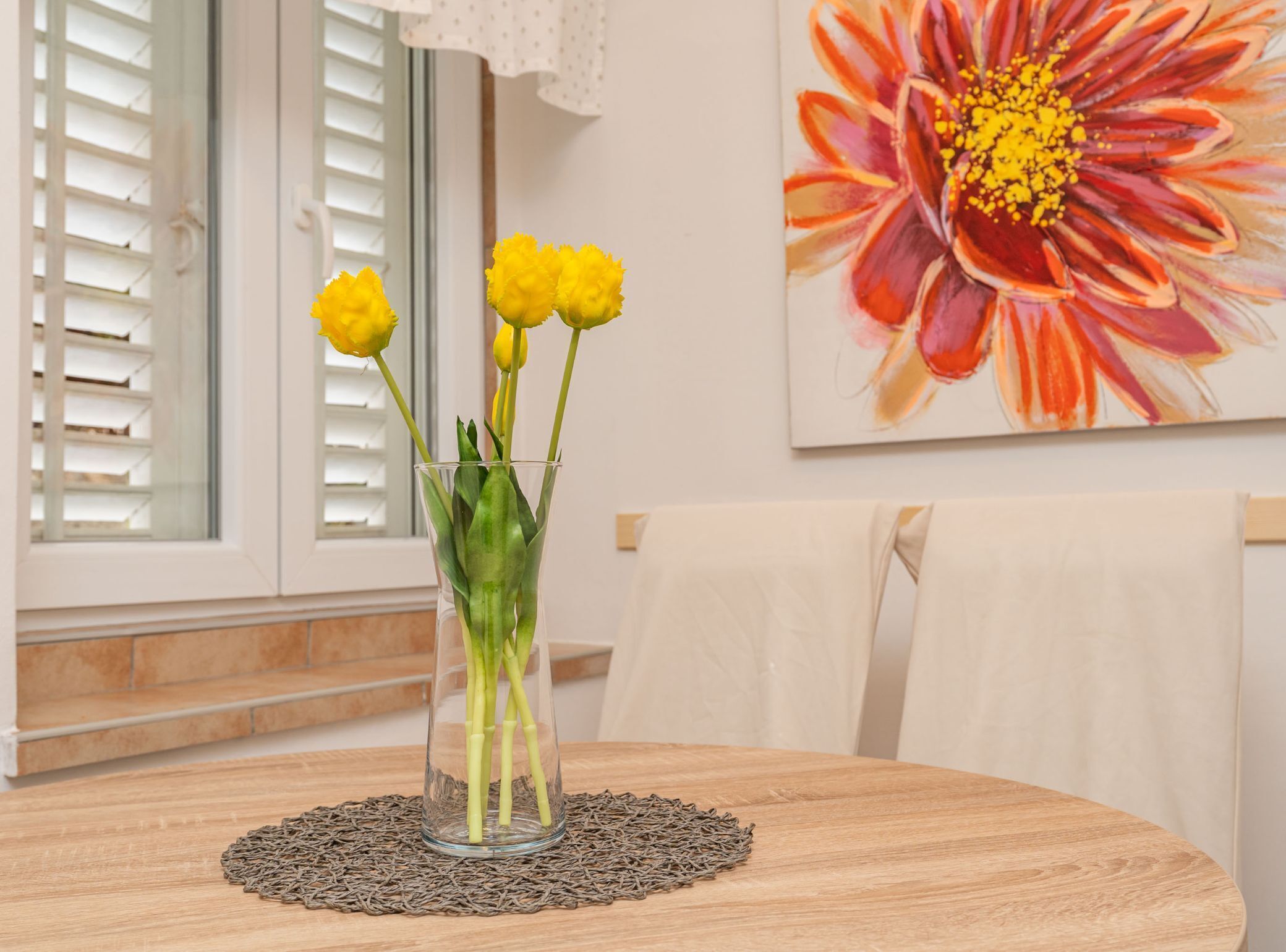 En gennemsigtig glasvase med gule blomster på et træbord, et vindue med hvide persienner, og et blomsterbillede på væggen.