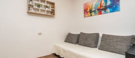 Un pequeño espacio de estar con un sofá, una estantería con plantas y un cuadro de colores en la pared.