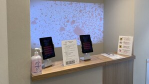 A reception desk with a digital screen displaying a welcome message, a sign for Sakura Cross Hotel, a hand sanitizer dispenser, and a caution sign.