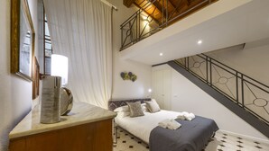 Ein Schlafzimmer mit Bett, Nachttisch, Spiegel, einem Fenster mit Vorhängen, einer Treppe und einer Wand mit dekorativen Tellern.