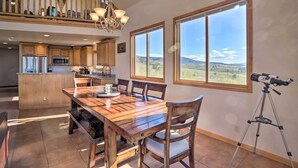 A spacious dining area with a wooden table and chairs, a kitchen in the background, and large windows offering a mountain view.