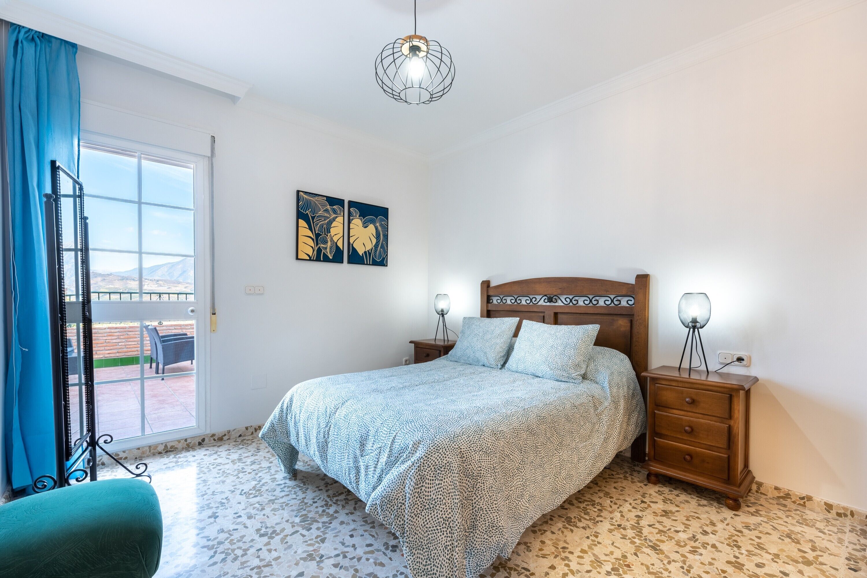Un dormitorio con cama, mesita de noche, lámpara, silla, ventana con cortinas y una obra de arte en la pared.