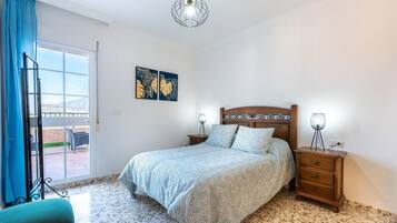 Un dormitorio con cama, mesita de noche, lámpara, silla, ventana con cortinas y una obra de arte en la pared.