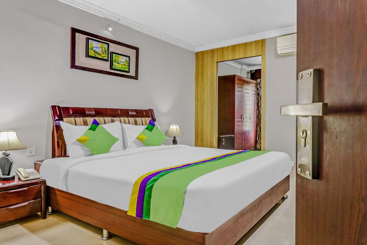 A hotel room with a wooden bed, white bedding, a green and purple striped throw, a bedside table with a lamp, and a phone. There are two framed pictures on the wall above the bed.