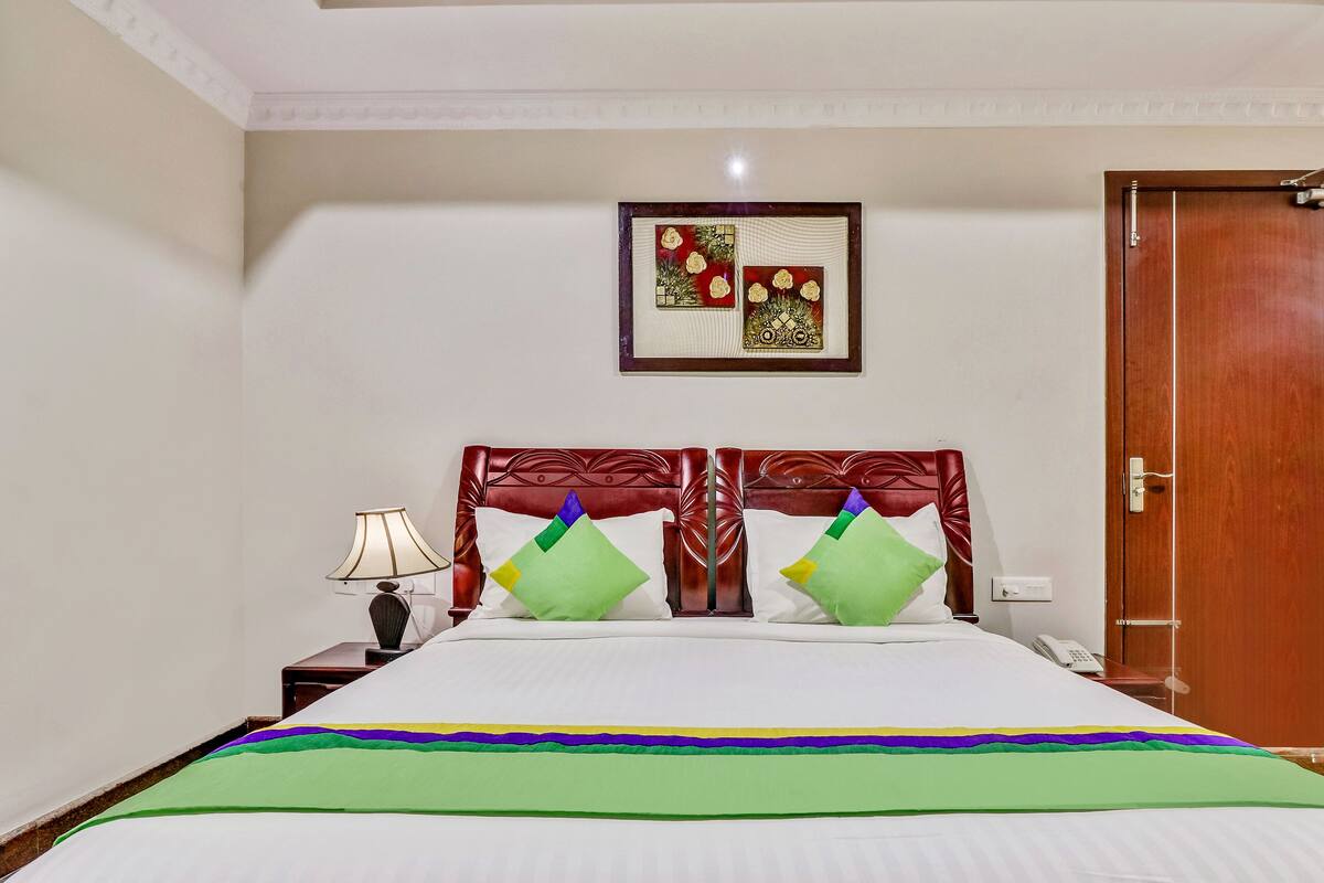 A hotel room with a bed, a nightstand, a lamp, a framed picture on the wall, and a wooden door.