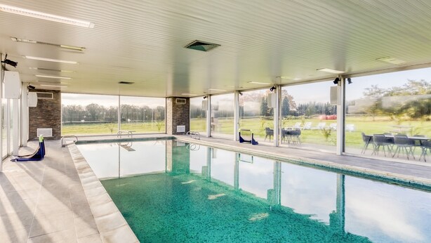 En modern inomhuspool med väggar av genomskinligt glas, en dekorativ vägg av sten och utsikt över en golfbana.