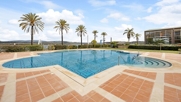 Une piscine dont le fond est recouvert de carreaux formant un motif géométrique, entourée de palmiers et surplombée d’un ciel dégagé.