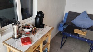 A small, tidy room with a window, a coffee maker, a bowl of snacks, a box of chocolates, and a blue sofa with cushions.