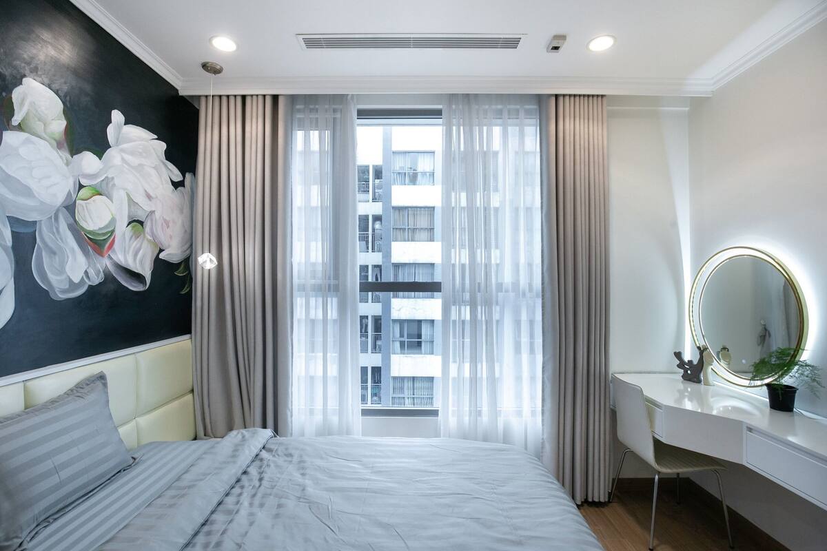 Ein modernes Hotelzimmer mit einem großen Bett, einem Schreibtisch mit Spiegel und Blick auf ein Gebäude durch leichte Vorhänge.