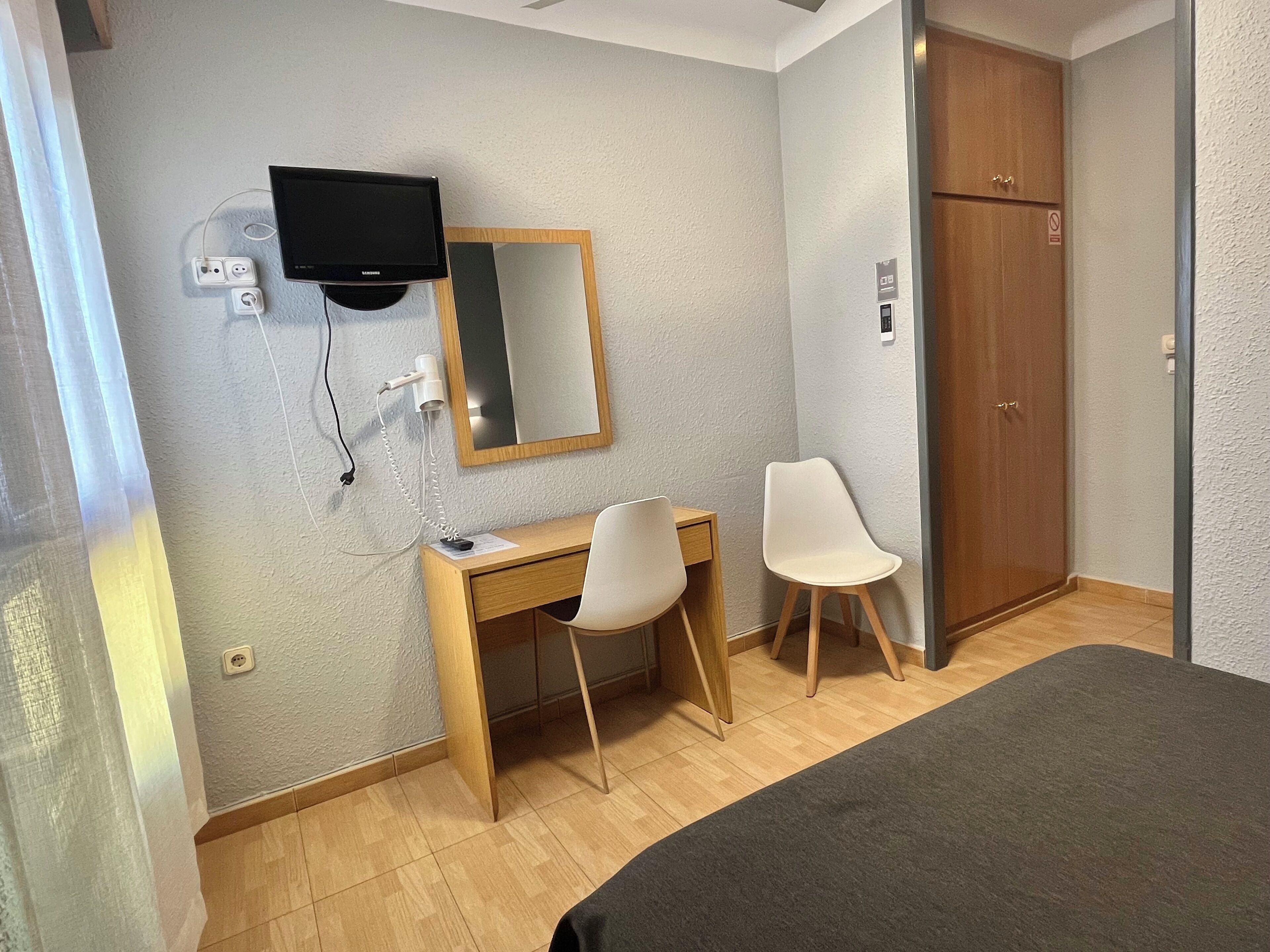 A hotel room with a wooden desk, two chairs, a television mounted on the wall, and a wardrobe.