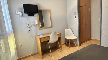 A hotel room with a wooden desk, two chairs, a television mounted on the wall, and a wardrobe.