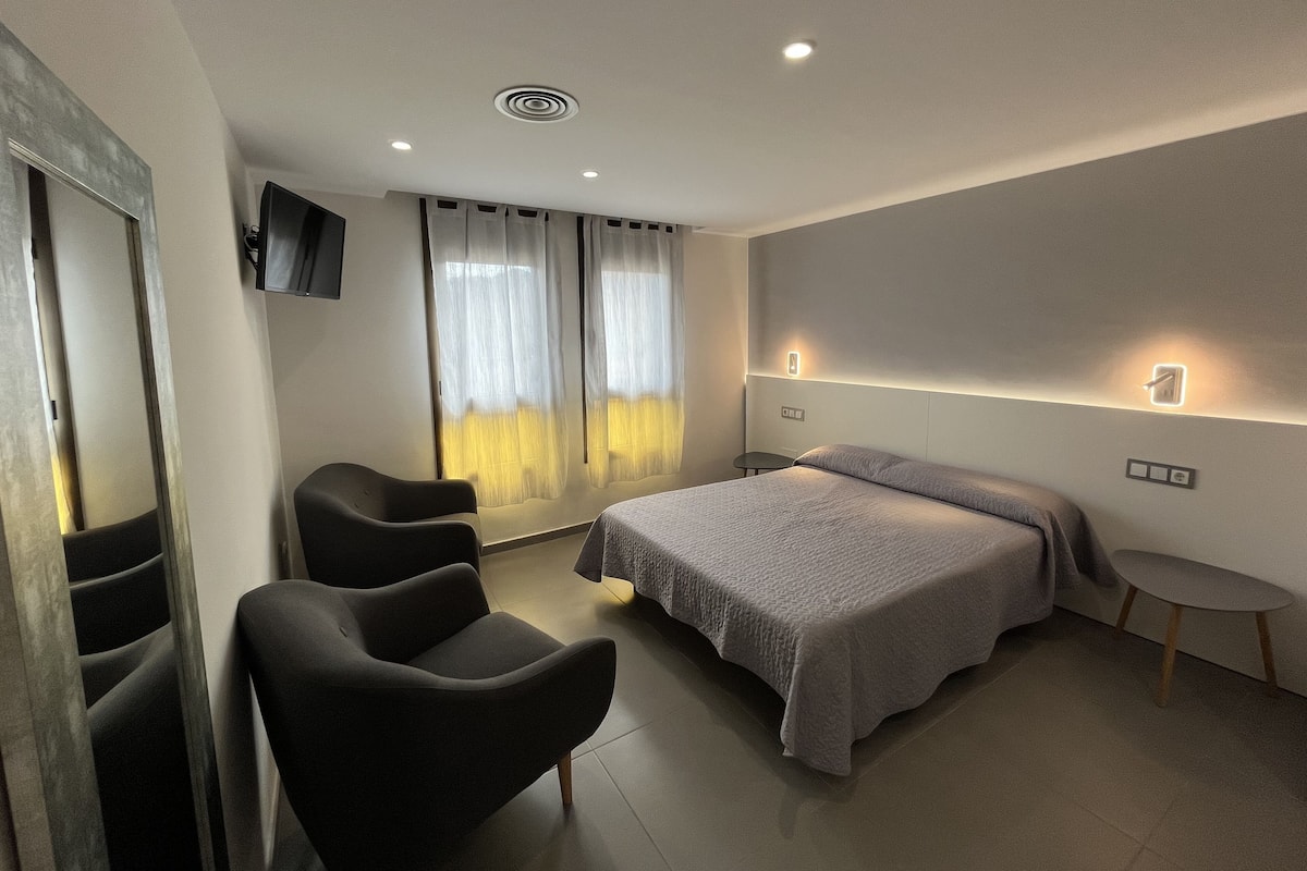 Una habitación de hotel moderna con cama, dos sillas, una mesita, televisión y ventana con cortinas.