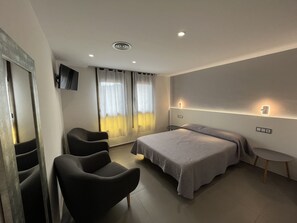 Een moderne hotelkamer met een bed, twee stoelen, een tafeltje, een televisie en een raam met gordijnen.