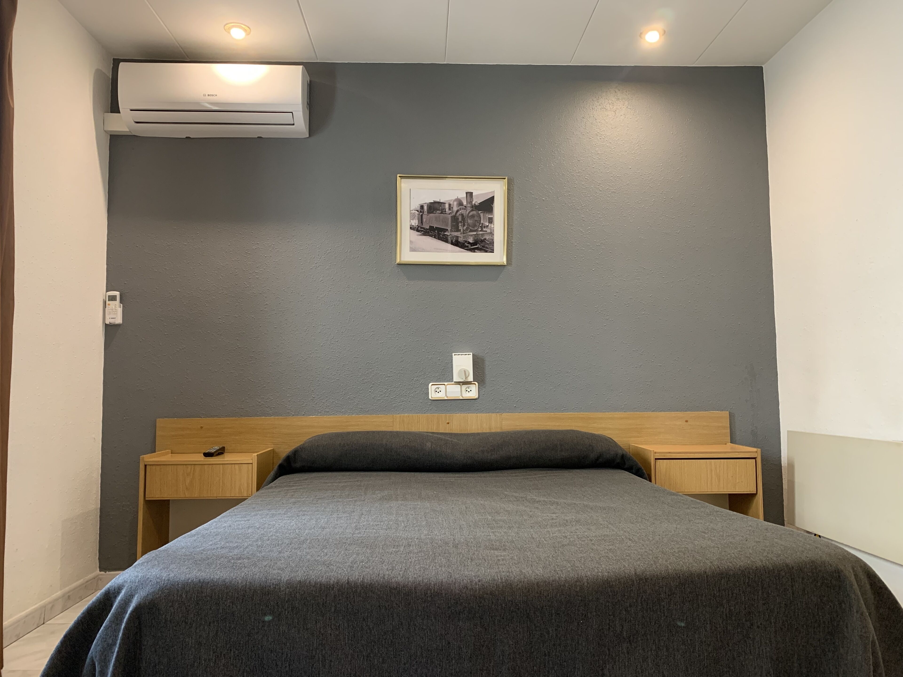 A bedroom with a grey bedspread, a wooden headboard, two bedside tables, a mounted air conditioner, a framed picture on the wall, and a power outlet.