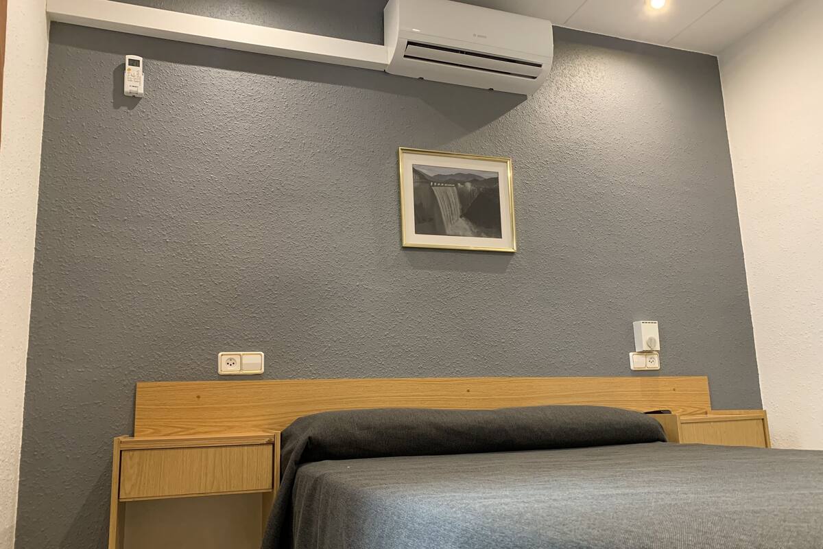 Un dormitorio con pared gris texturizada, un cuadro enmarco, cabecera de madera, cama con manta gris y un equipo de aire acondicionado.