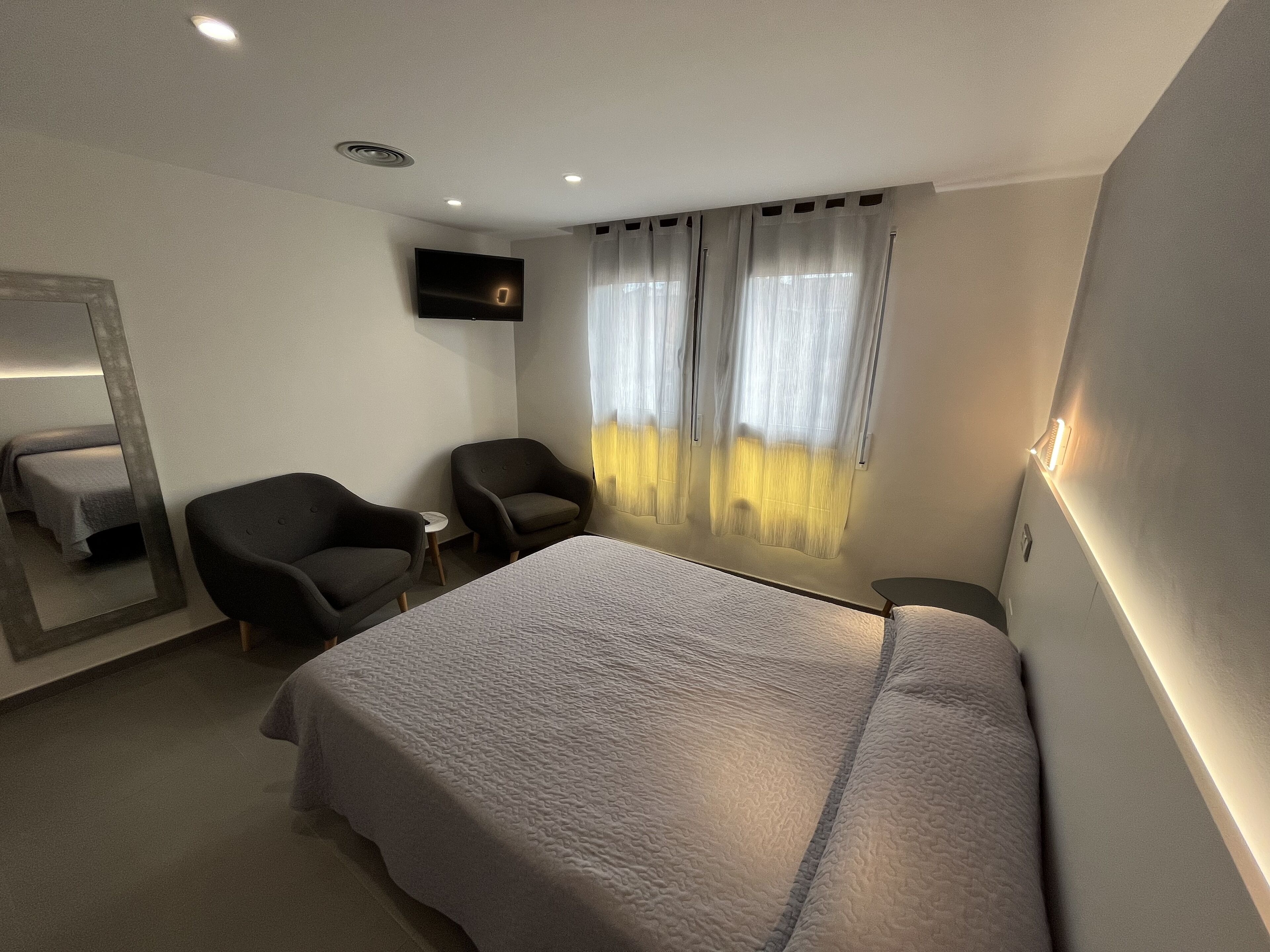 Habitación de hotel con una cama grande, dos sillones, una mesita, un televisor montado en la pared y una ventana con cortinas translúcidas.