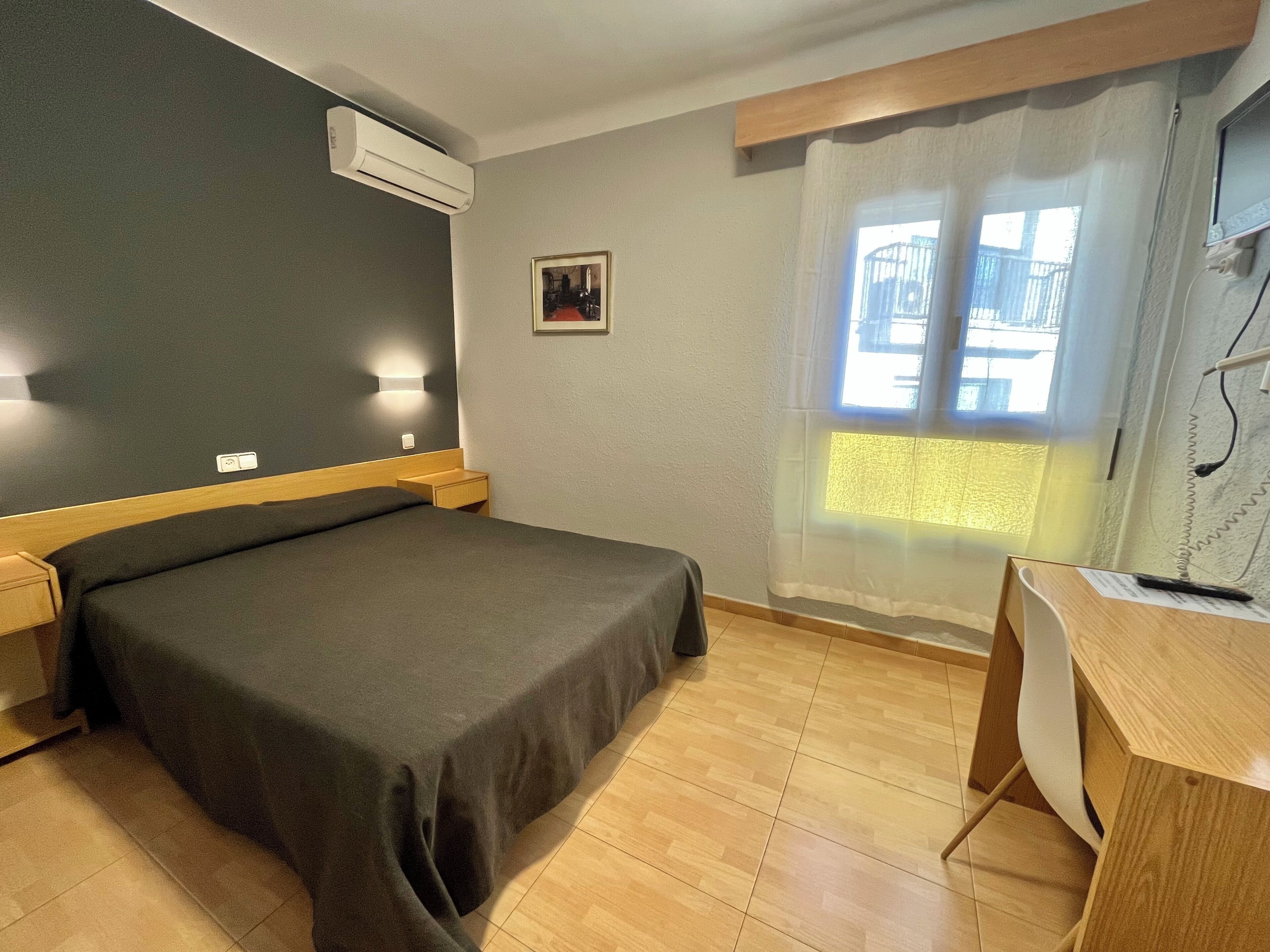 Een hotelkamer met een bed, nachtkastjes, een airconditioningapparaat, een bureau met stoel, een raam met gordijnen, en een klein schilderij aan de muur.