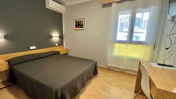 Een hotelkamer met een bed, nachtkastjes, een airconditioningapparaat, een bureau met stoel, een raam met gordijnen, en een klein schilderij aan de muur.