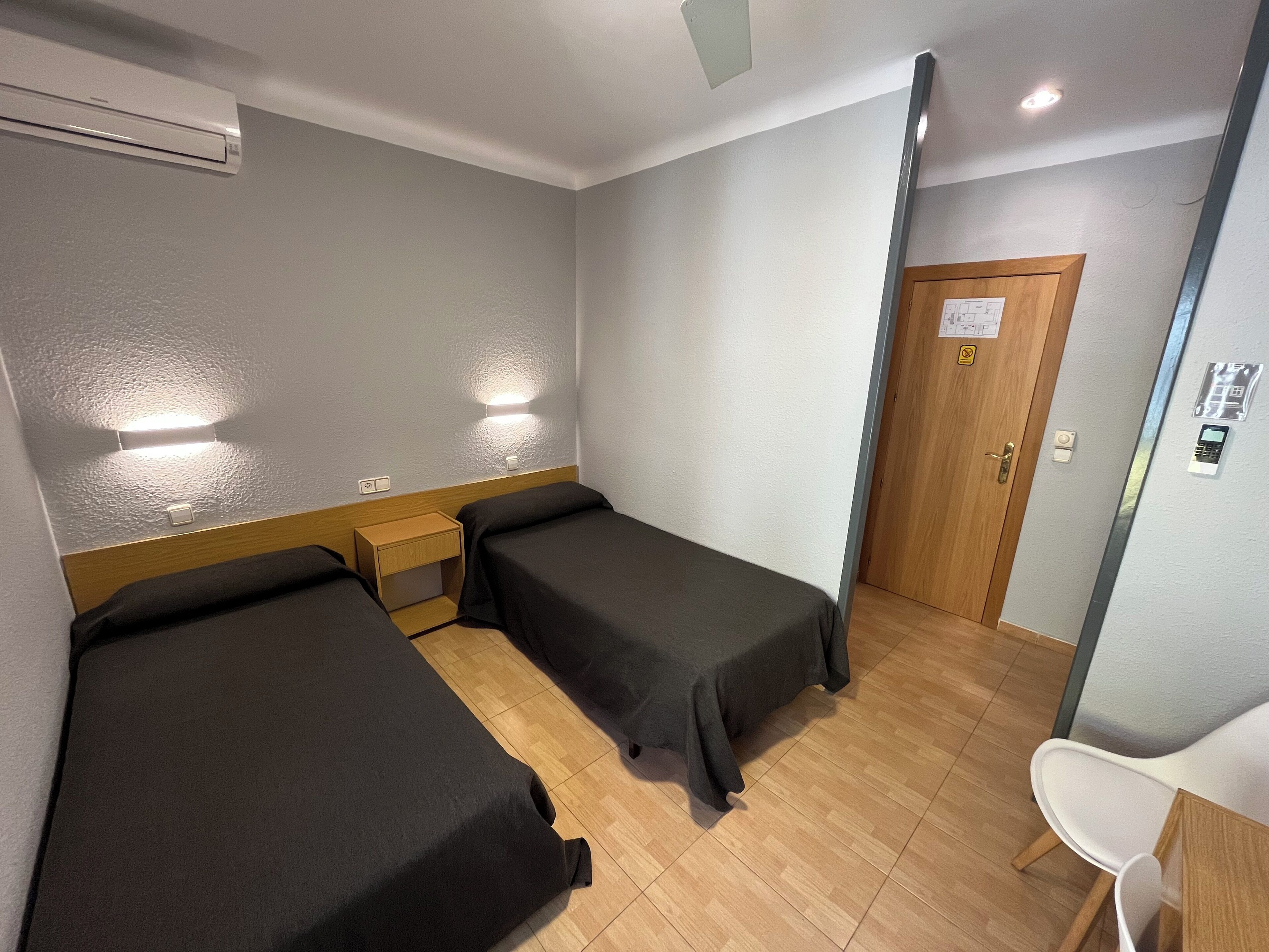 Hotelkamer met twee eenpersoonsbedden, een houten nachtkastje, een bureau met stoel, een airconditioning en een wandlamp. De deur heeft een codeerapparaat.