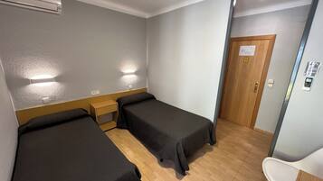 Hotelkamer met twee eenpersoonsbedden, een houten nachtkastje, een bureau met stoel, een airconditioning en een wandlamp. De deur heeft een codeerapparaat.