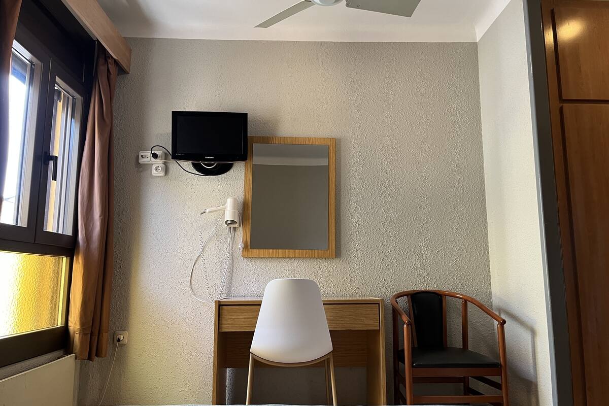 Habitación de hotel con cama, escritorio y silla, televisión, ventilador de techo y ventana con cortinas.